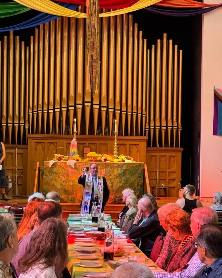 Photo of Rev. Mike Mather holding the communion elements at the head of a lunch table in the sanctuary.