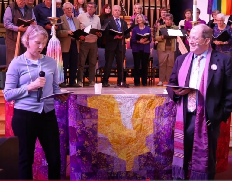 Pastor Mike and Lexi Fortna in front of the altar, with the Chancel Choir in the background.