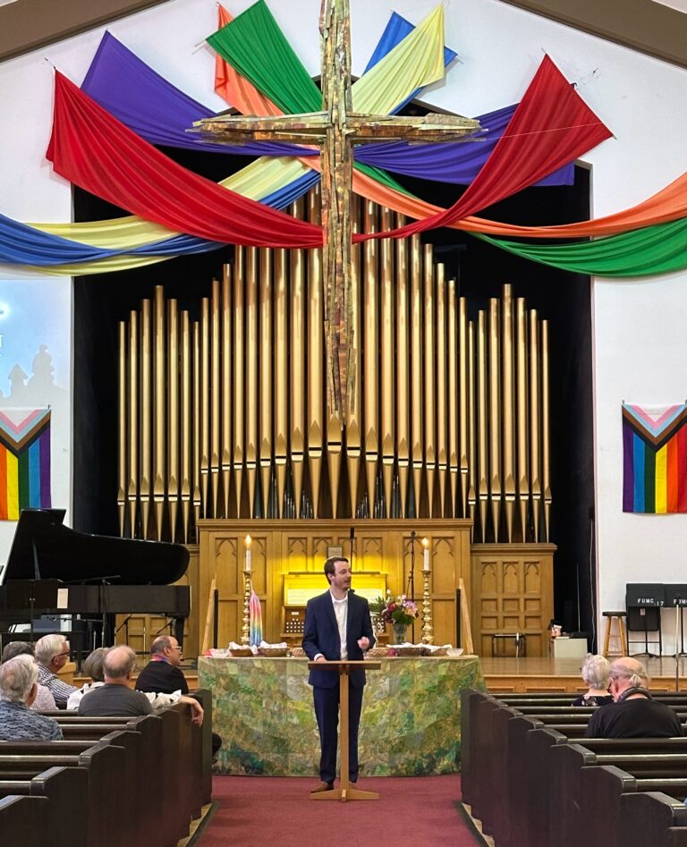Kevin Lazarus preaching in front of the altar, cross, and rainbow swaggeries