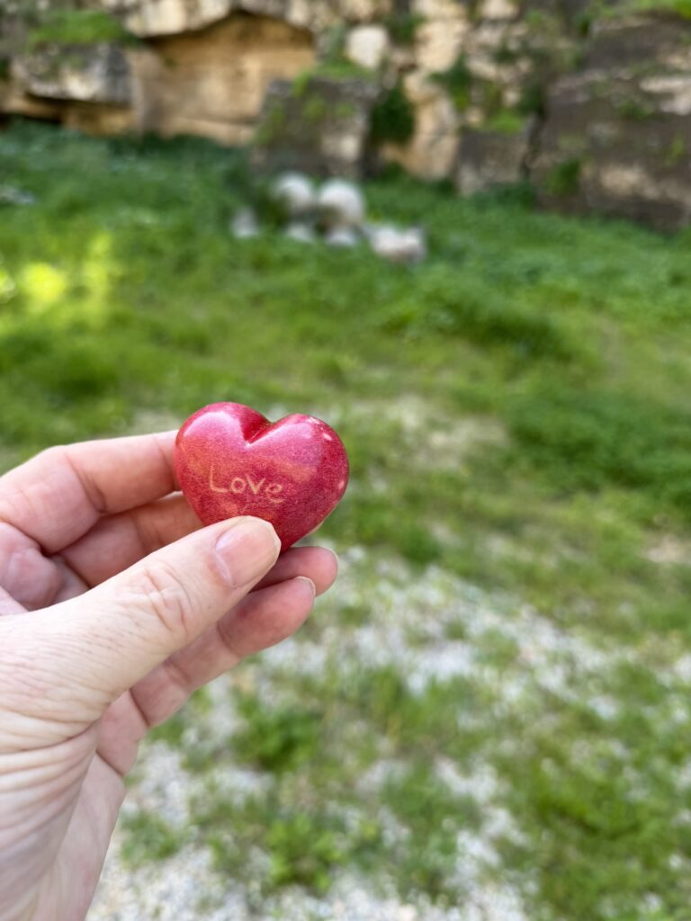 Pastor Mike's hand holding a polished, red heart-shaped rock engraved with "love" in front of cannonballs in Rhodes, Greece